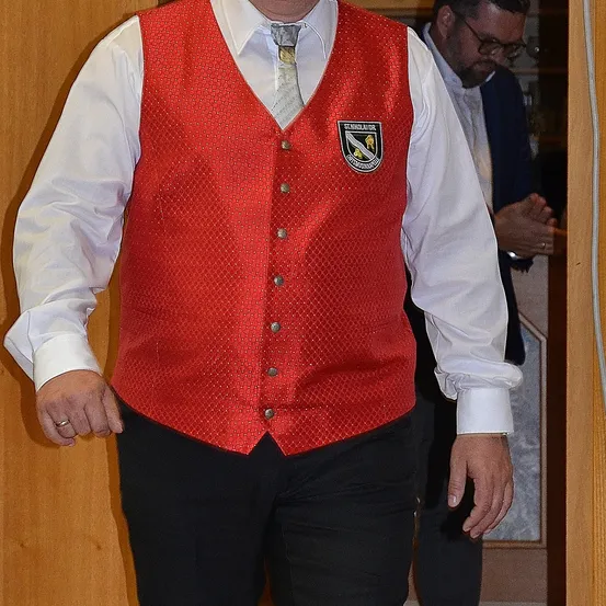 Ein Mann in einem roten Sakko mit Logo steht neben einem Mann in einem blauen Anzug und mit Brille.