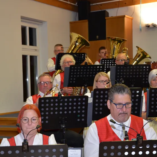 Eine Band von Musikern, einschließlich Blech- und Holzblasinstrumente, spielt auf der Bühne. Sie tragen rote Westen und einige tragen Brillen. Es gibt Notenständer vor ihnen.