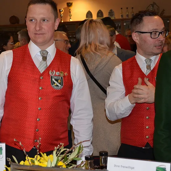 Zwei Männer in roten Westen mit einem Logo stehen inmitten einer Menschenmenge bei einer Veranstaltung in Innenräumen, einer berührt seine Brust. Hinter ihnen eine Frau mit langen blonden Haaren und ein Mann mit Brille. Ein Tisch mit einem Schild und Blumen ist im Vordergrund.
