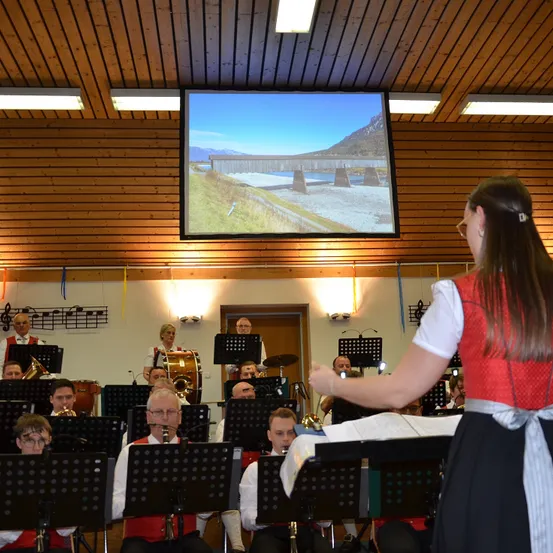 Ein Orchester spielt Instrumente, während eine Frau dirigiert. Eine Projektionsleinwand zeigt eine Landschaft. Der Raum hat Holzwände und Deckenleuchten.