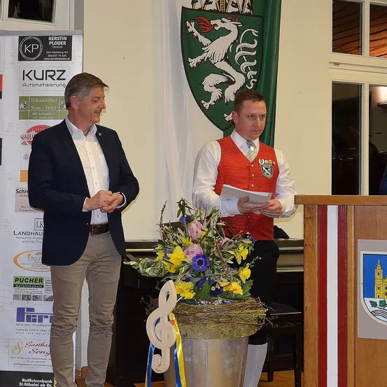 Zwei Männer in formeller Kleidung stehen neben einem Podium. Ein Mann liest aus einem Stück Papier, während der andere zusieht. Ein Blumenarrangement und ein Banner mit einem Wappen im Hintergrund sind zu sehen.