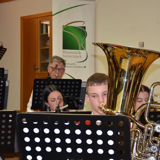 Eine Gruppe von Musikern in einem Musikraum spielt Instrumente, einschließlich einer Tuba, während ein Banner mit dem Text Blasmusik Steiermark hinter ihnen ist.
