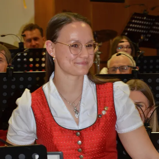 Eine Frau in einem roten Kleid mit weißen Rändern sitzt unter anderen Musikern und lächelt für die Kamera. Hinter ihr zeigt ein verschwommener Hintergrund andere Personen mit Instrumenten und Notenständern.