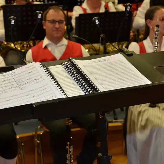 Ein Orchester probet, mit Noten und Instrumenten sichtbar. Ein Mann und eine Frau halten Klarinetten, während ein Mann in einem roten Westchen Musik liest.