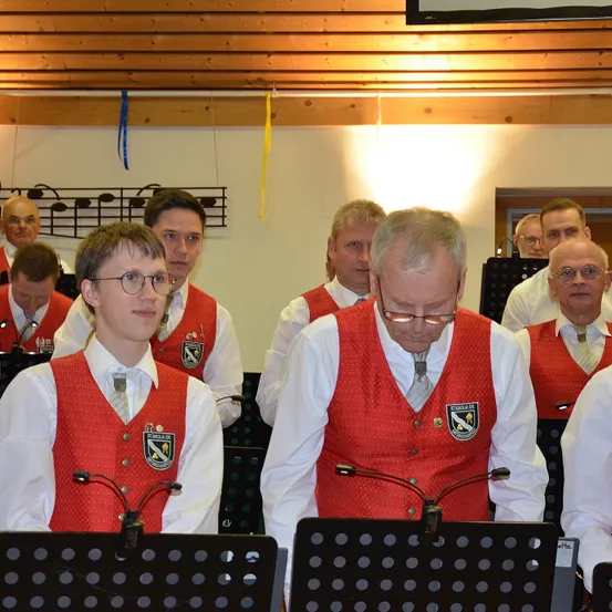 Ein Männerchor in roten Westen mit weißen Hemden und Krawatten singt ein Lied. Sie stehen vor Notenständern mit Mikrofonen.