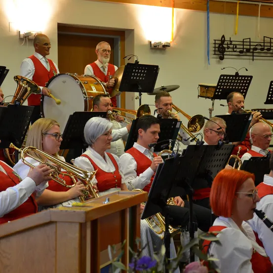 Ein Blasorchester spielt in einem Saal mit mehreren Musikern. Sie tragen rote Westen. Eine Trommel steht vorne, und verschiedene Blechblasinstrumente dahinter. Musiknoten hängen an der Wand.