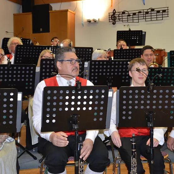 Eine Gruppe von Musikern in traditioneller Kleidung sitzt in einer Reihe mit ihren Instrumenten, darunter Klarinetten und Saxophone, in einem Raum mit Noten an der Wand.