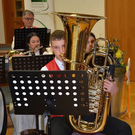 Ein junger Mann spielt Tuba in einer Band mit anderen Musikern, darunter eine Frau, die Flöte spielt, während er in einem Raum mit Holzwänden sitzt.