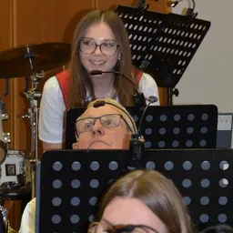 Eine Frau mit Brille singt in ein Mikrofon, während ein Mann mit Kopfhörern zuhört. Dahinter ist das Drum-Setup eines Schlagzeugers mit Becken zu sehen.