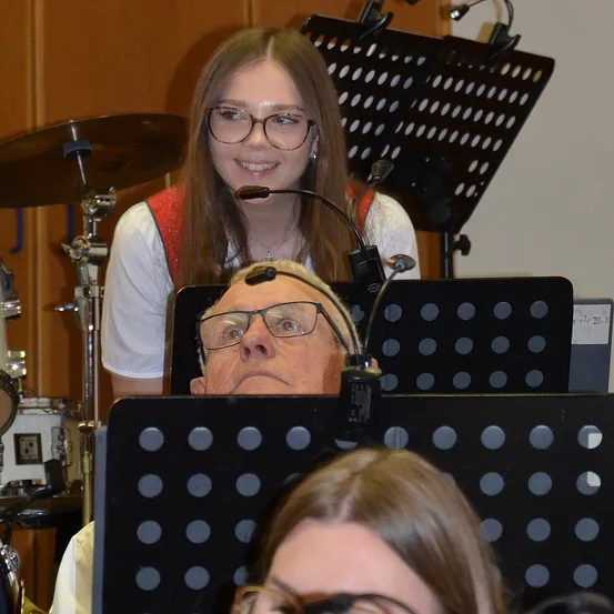 Eine Frau mit Brille singt in ein Mikrofon, während ein Mann mit Kopfhörern zuhört. Dahinter ist das Drum-Setup eines Schlagzeugers mit Becken zu sehen.