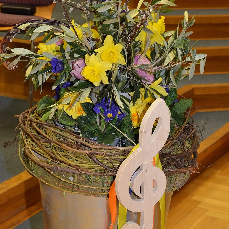Ein Korb mit leuchtenden Blumen, darunter gelbe Narzissen und lila Blüten, steht auf einem Holzboden mit einer musikalischen Noten-Dekoration.