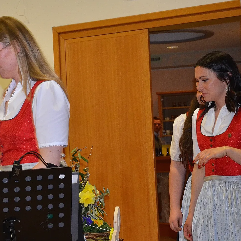 Zwei Frauen stehen in einem Raum, eine schaut nach vorne, die andere zur Seite. Beide tragen traditionelle deutsche Kleidung. Ein Notenständer und Blumen sind in der Nähe.