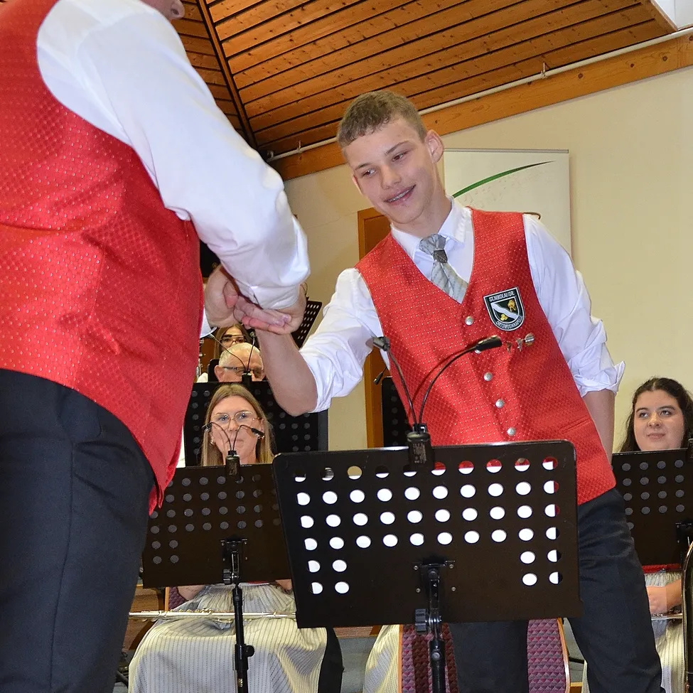 Ein junger Mann in einer roten Weste und Krawatte gibt einem Erwachsenen in einer roten Weste in einer Kirche die Hand. Dahinter sitzt eine Frau auf einem Stuhl mit einem Notenständer.