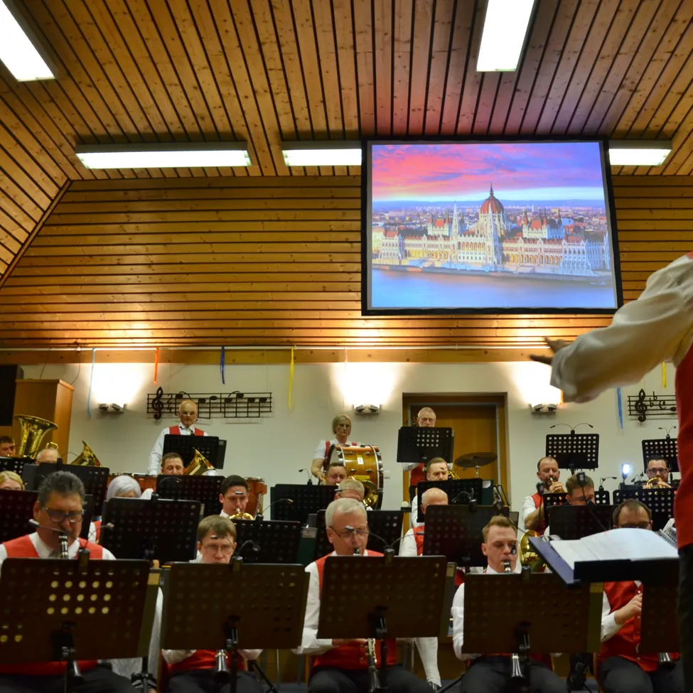 Ein großes Orchester spielt in einem Saal, wobei eine Projektionsleinwand ein Bild des ungarischen Parlaments in Budapest zeigt.
