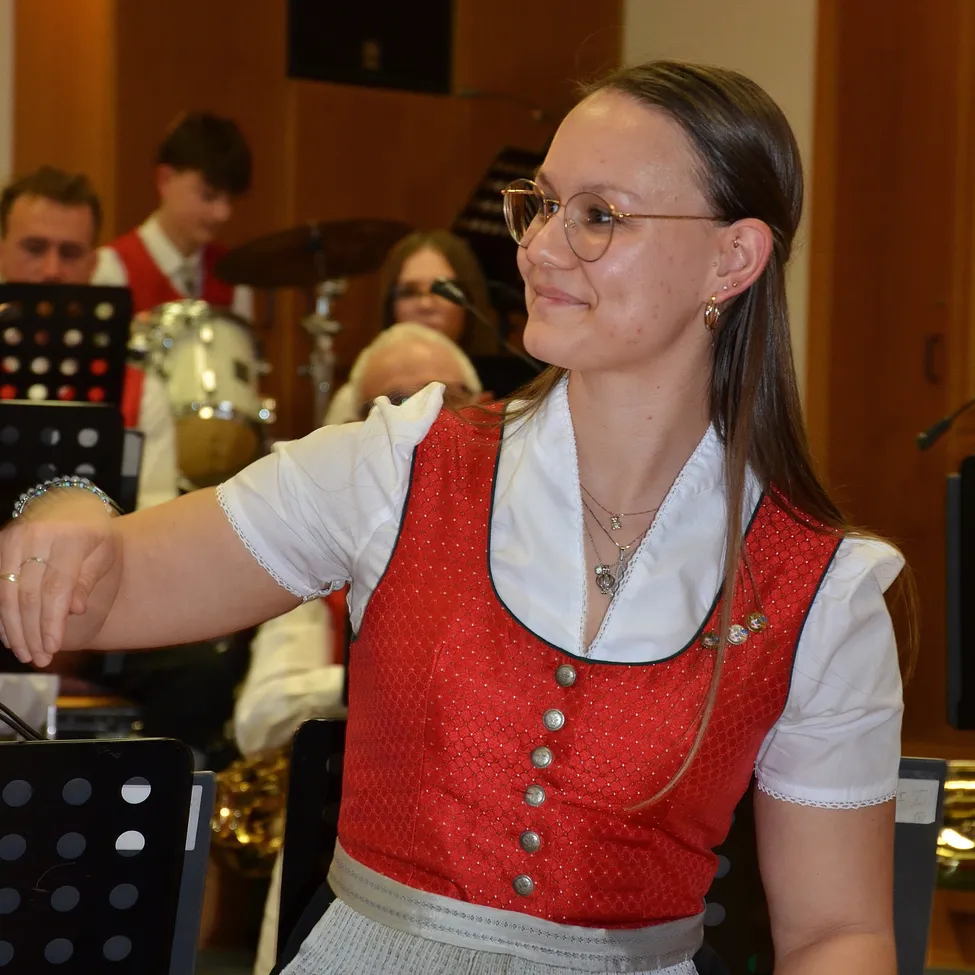 Eine Frau in einem roten und weißen traditionellen Kleid steht, lächelt und hat ihre Hand auf der Schulter von jemandem. Hinter ihr übt eine Band mit verschiedenen Instrumenten.