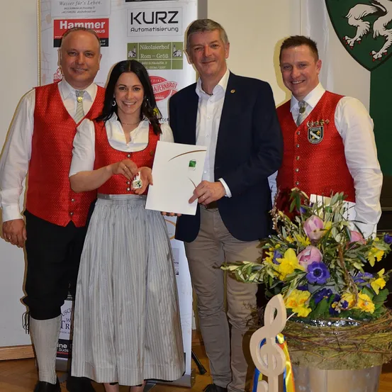 Eine Frau hält eine Auszeichnung in der Hand, während sie neben drei Männern in traditioneller Kleidung steht. Hinter ihnen befindet sich ein Banner mit verschiedenen Logos und einem Wappen.