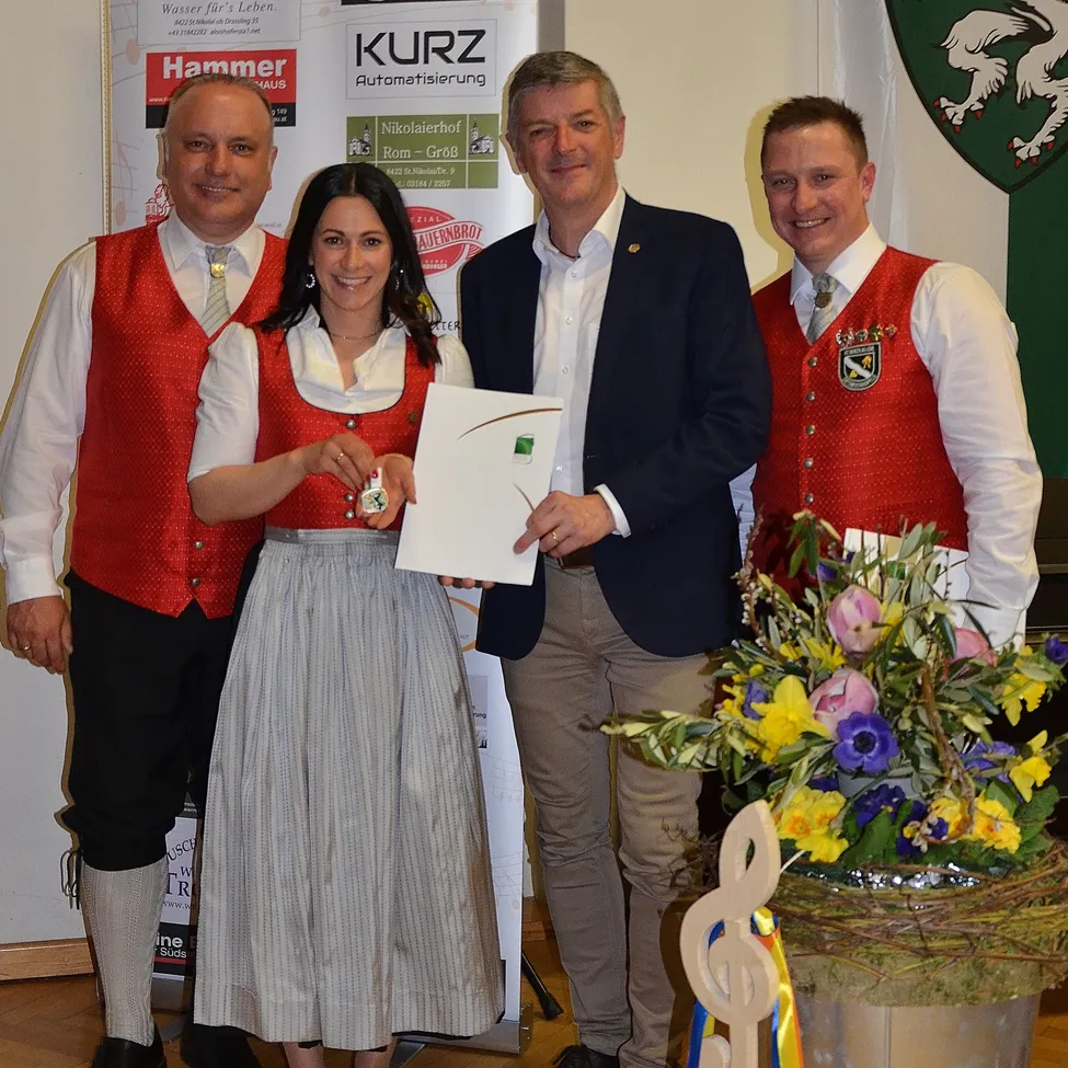 Eine Frau hält eine Auszeichnung in der Hand, während sie neben drei Männern in traditioneller Kleidung steht. Hinter ihnen befindet sich ein Banner mit verschiedenen Logos und einem Wappen.