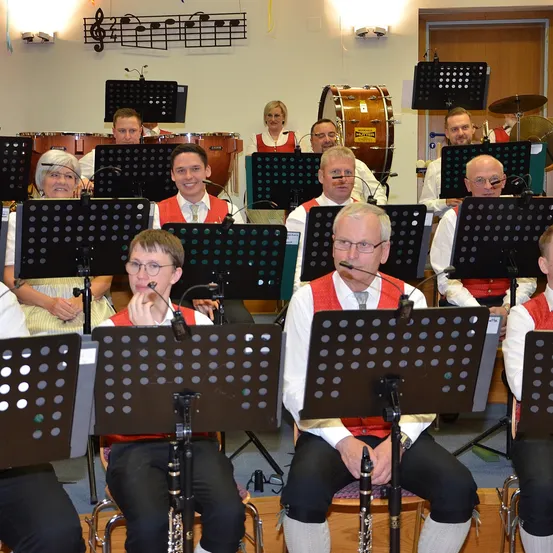 Eine Gruppe von Musikern in roten Westen sitzen und stehen auf Stühlen und spielen verschiedene Instrumente. Noten und Instrumente hängen an der Wand.
