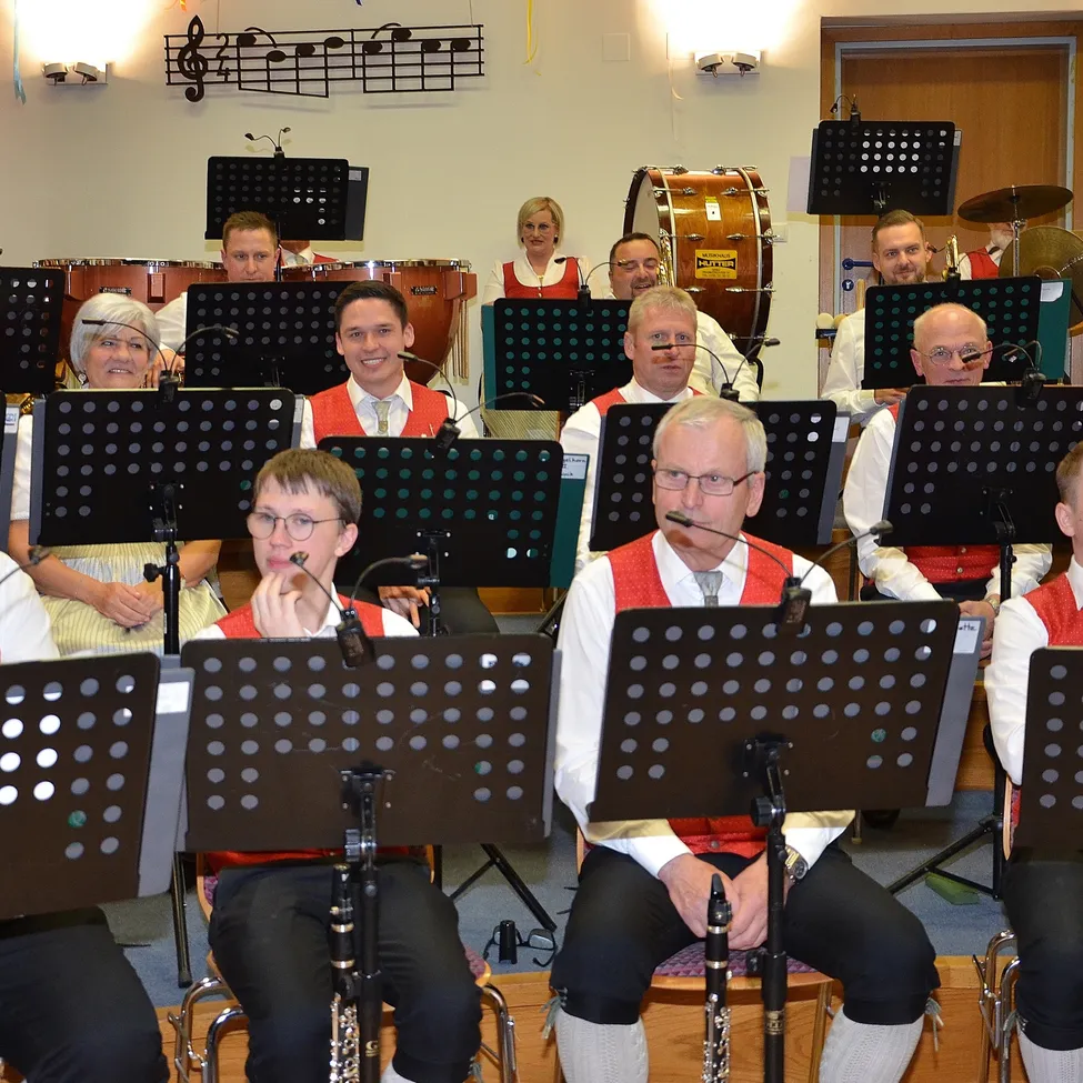 Eine Gruppe von Musikern in roten Westen sitzen und stehen auf Stühlen und spielen verschiedene Instrumente. Noten und Instrumente hängen an der Wand.