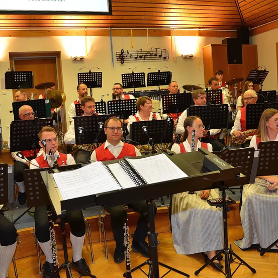 Eine Gruppe von Musikern, einschließlich Blech- und Holzblasinstrumenten, versammelt sich in einem Saal, möglicherweise für eine Aufführung, mit Notenständern, Mikrofonen und Musikinstrumenten vor ihnen.
