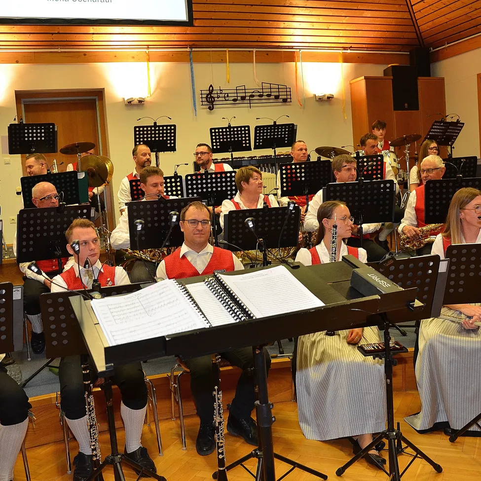 Eine Gruppe von Musikern, einschließlich Blech- und Holzblasinstrumenten, versammelt sich in einem Saal, möglicherweise für eine Aufführung, mit Notenständern, Mikrofonen und Musikinstrumenten vor ihnen.