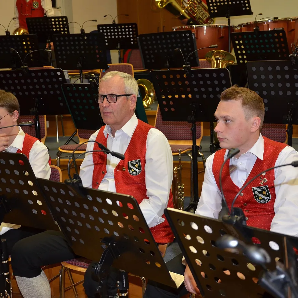 Drei Männer in roten Westen sitzen an Notenpulten mit Instrumenten dahinter, einer mit Brille, ein anderer mit Mikrofon.