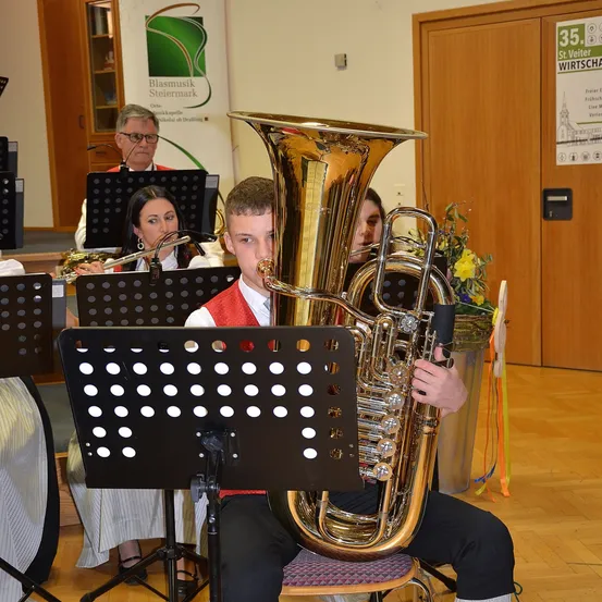 Ein junger Mann spielt eine große Messingtuba, während er auf einem Stuhl sitzt, umgeben von einer kleinen Band. Hinter ihm spielt eine Frau Flöte, und ein Mann spielt Saxophon. Sie sind in einem Raum mit Holzwänden und einem Holzboden.