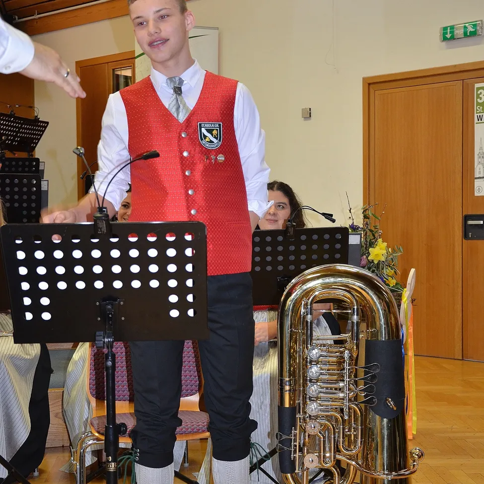 Ein junger Mann mit einem roten Westchen steht vor einer Tuba und hält einen Notenständer mit einem Mikrofon. Er befindet sich in einem Raum mit Holztüren und anderen Musikinstrumenten im Hintergrund.