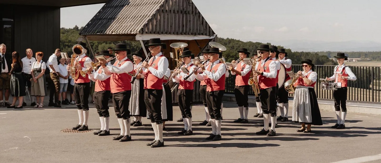 Eine Gruppe von Musikern in traditioneller Kleidung tritt im Freien auf, steht in einer Reihe mit Instrumenten. Dahinter ist ein Gebäude mit einem dreieckigen Dach zu sehen, und Bäume sind im Hintergrund.