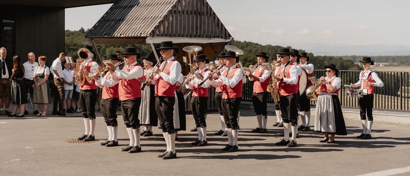 Eine Gruppe von Musikern in traditioneller Kleidung tritt im Freien auf, steht in einer Reihe mit Instrumenten. Dahinter ist ein Gebäude mit einem dreieckigen Dach zu sehen, und Bäume sind im Hintergrund.