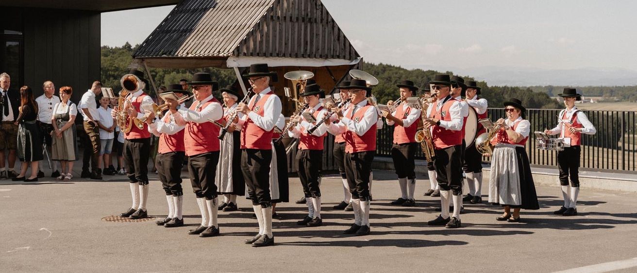 Eine Gruppe von Musikern in traditioneller Kleidung tritt im Freien auf, steht in einer Reihe mit Instrumenten. Dahinter ist ein Gebäude mit einem dreieckigen Dach zu sehen, und Bäume sind im Hintergrund.
