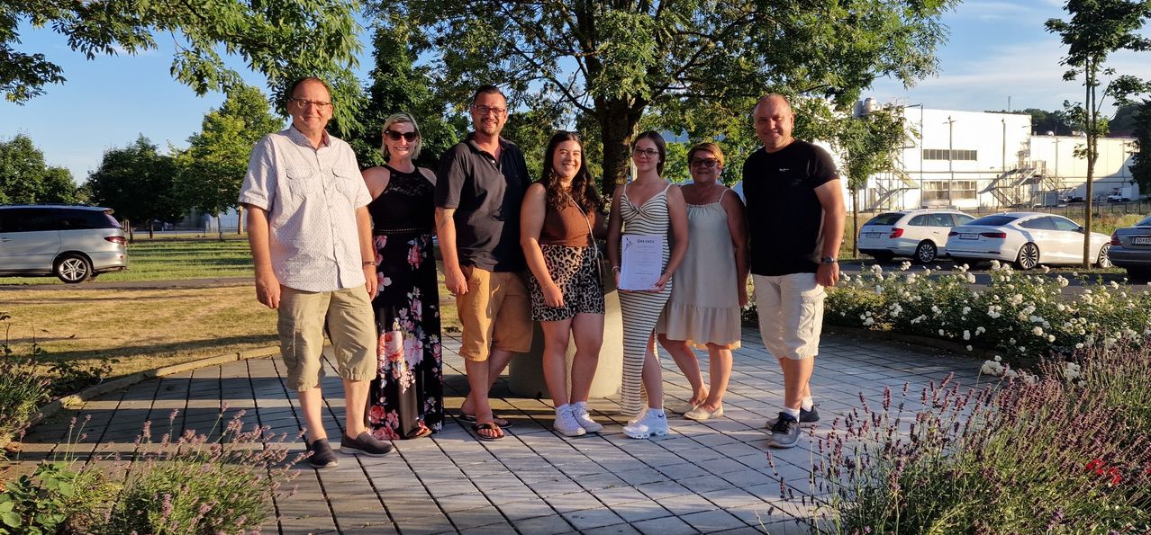 Eine Gruppe von sieben Personen posiert für ein Foto im Freien. Eine Frau hält ein Zertifikat. Bäume und ein gepflasterter Weg sind im Hintergrund zu sehen.