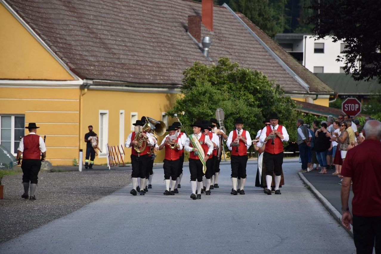 Eine Blaskapelle spielt Instrumente, während sie auf der Straße geht. Einige spielen Trompeten, andere spielen Tuben. Dahinter stehen Gebäude und Bäume.