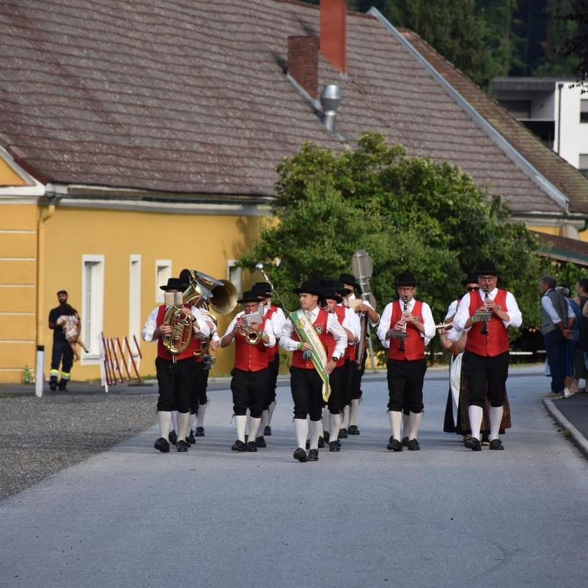 Eine Blaskapelle spielt Instrumente, während sie auf der Straße geht. Einige spielen Trompeten, andere spielen Tuben. Dahinter stehen Gebäude und Bäume.