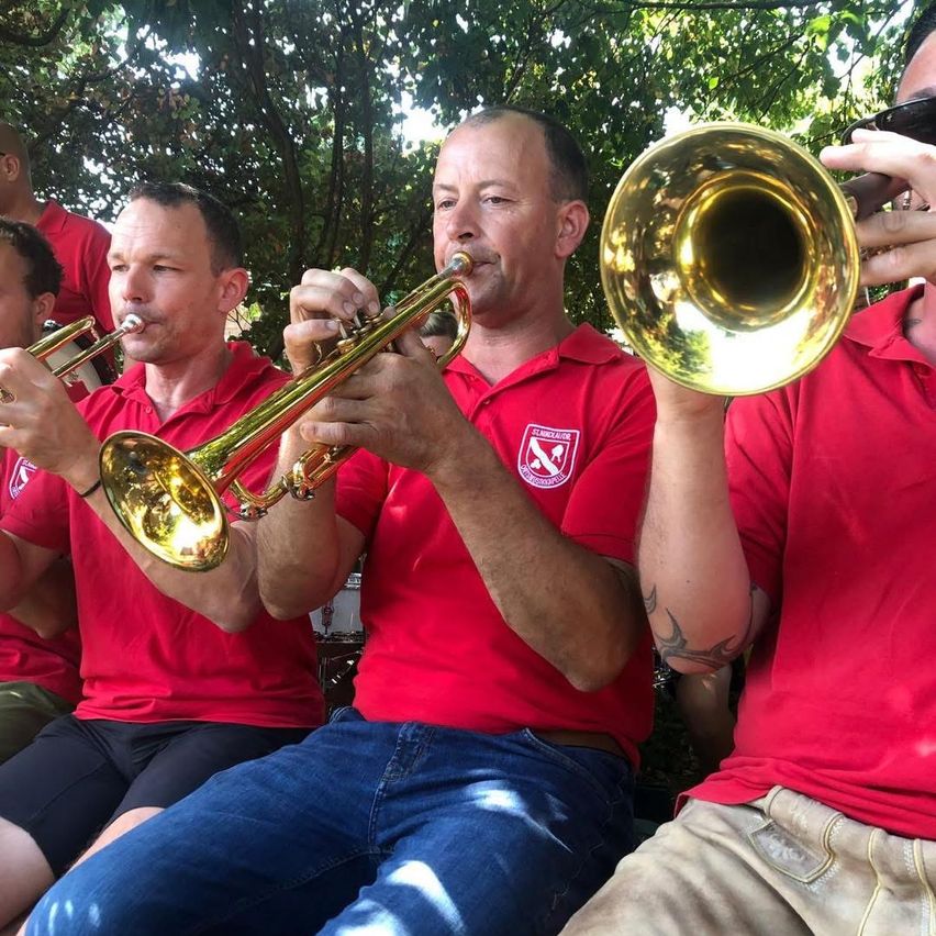 Vier Männer in roten Shirts spielen Trompeten im Freien. Einer hält eine Trompete in jeder Hand, während die anderen jeweils eine einzelne Trompete spielen. Sie sitzen vor einem Baum.