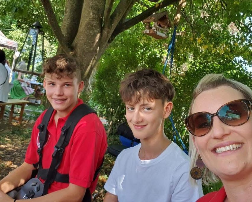 Drei junge Personen, zwei Jungen und ein Mädchen, lächeln für ein Foto unter einem Baum. Der Junge auf der linken Seite trägt ein rotes Hemd und einen Rucksack. Der mittlere Junge trägt ein weißes Hemd. Das Mädchen trägt eine Sonnenbrille und ein rotes Hemd.