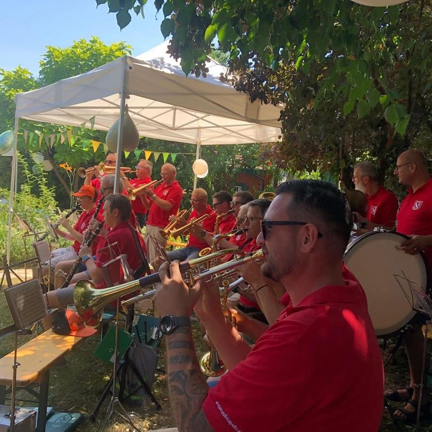 Eine Gruppe von Musikern in roten Shirts spielt verschiedene Instrumente im Freien unter einem weißen Zelt. Die Umgebung ist von Grün umgeben und umfasst eine Trommel und verschiedene Blasinstrumente.