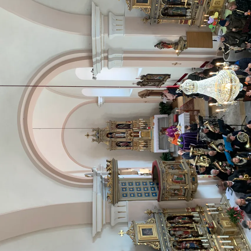 Der Innenraum einer Kathedrale mit einem großen Bogen, kunstvollen Altären und Musikern, die verschiedene Instrumente spielen. Eine große Kristallleuchter hängt von der Decke ab.