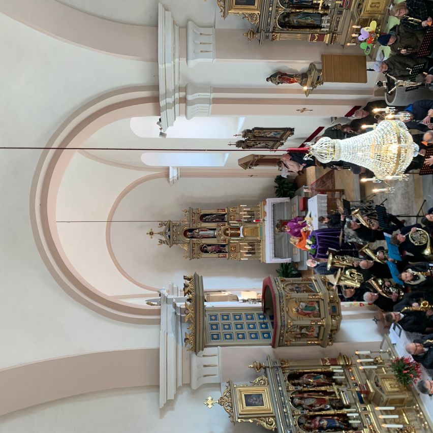 Der Innenraum einer Kathedrale mit einem großen Bogen, kunstvollen Altären und Musikern, die verschiedene Instrumente spielen. Eine große Kristallleuchter hängt von der Decke ab.