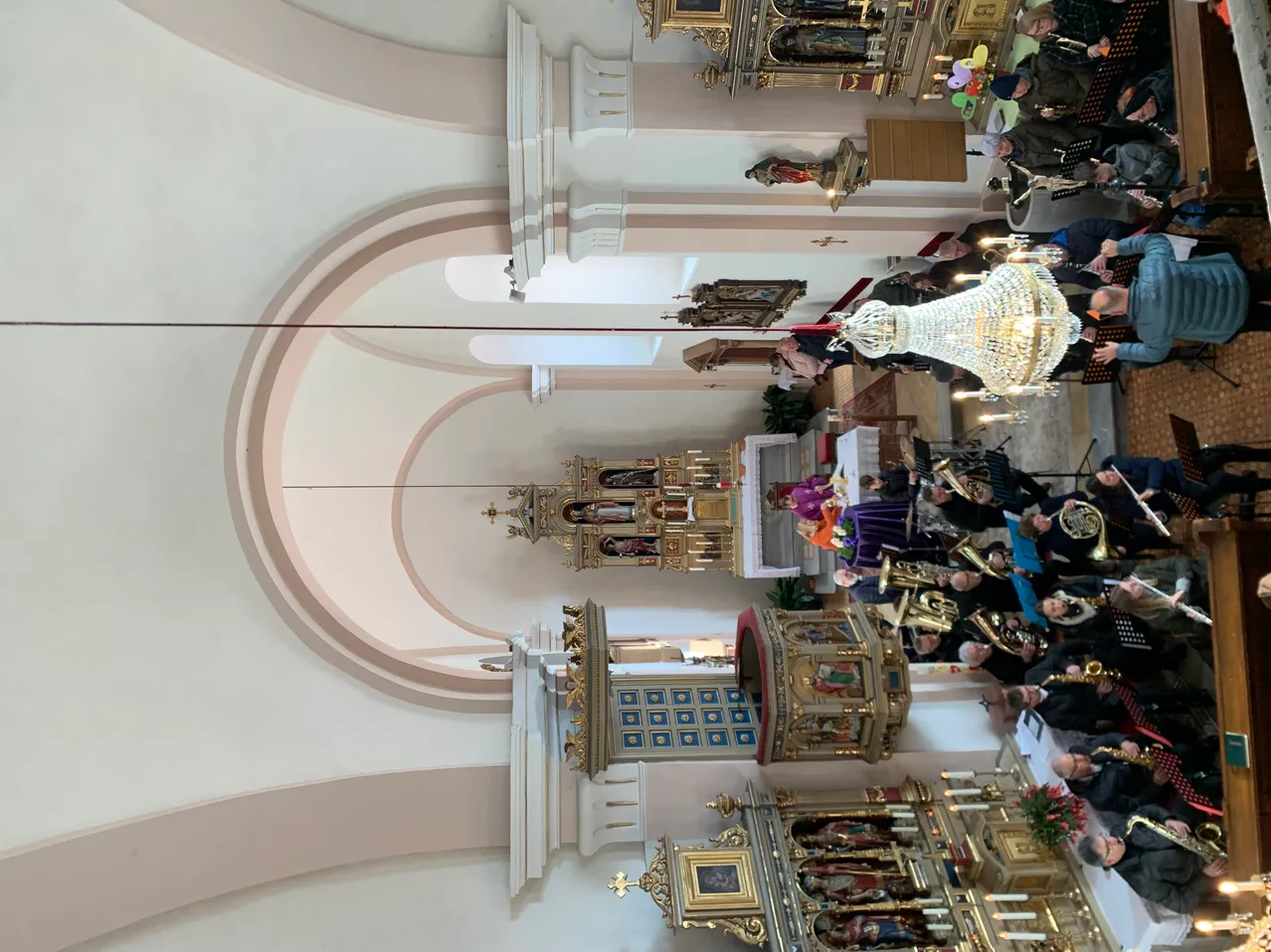 Der Innenraum einer Kathedrale mit einem großen Bogen, kunstvollen Altären und Musikern, die verschiedene Instrumente spielen. Eine große Kristallleuchter hängt von der Decke ab.