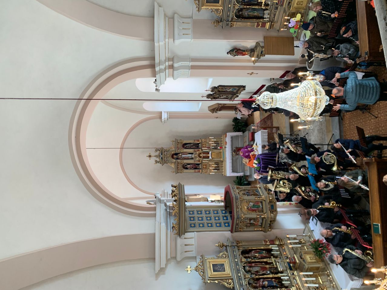 Der Innenraum einer Kathedrale mit einem großen Bogen, kunstvollen Altären und Musikern, die verschiedene Instrumente spielen. Eine große Kristallleuchter hängt von der Decke ab.