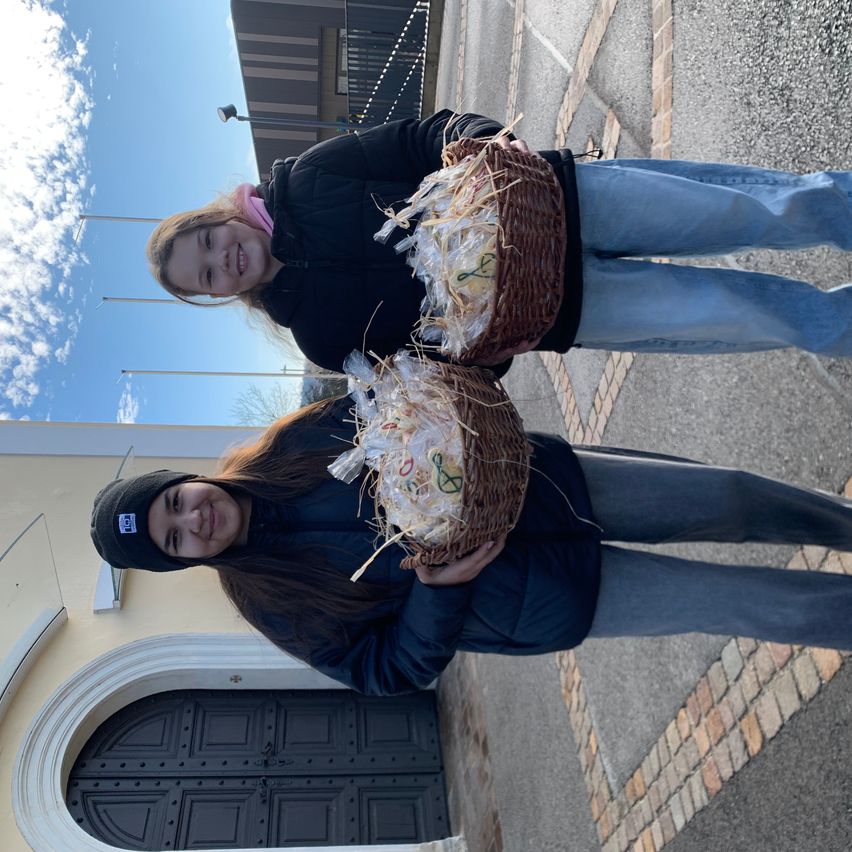 Zwei Frauen mit Körben voller Brot stehen vor einem Gebäude mit einem bogenförmigen Eingang und einem bewölkten Himmel darüber.