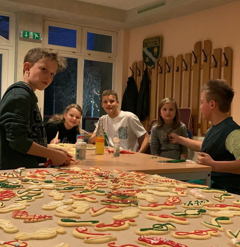Eine Gruppe von Kindern ist um einen Tisch versammelt, auf dem dekorierte Kekse stehen. Sie arbeiten an den Designs mit Zuckerguss. Der Raum ist warm beleuchtet und an der Wand befindet sich ein Schild mit einem Logo.