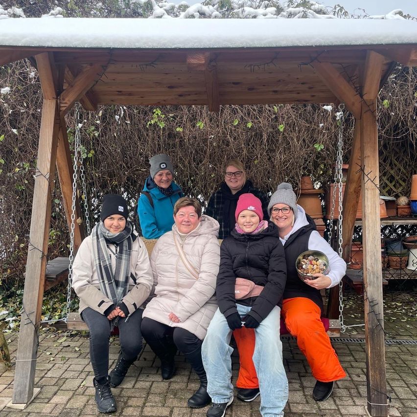 Sechs Frauen sitzen unter einem hölzernen Unterstand und lächeln für ein Foto. Sie tragen Winterkleidung. Eine Frau hält eine Schüssel mit Essen. Der Unterstand hat ein schneebedecktes Dach.