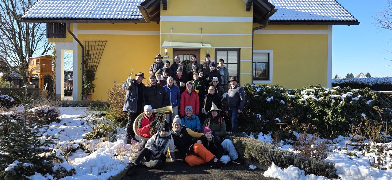Eine Gruppe von Menschen in Winterkleidung, einige mit Mützen und Brillen, stehen und sitzen vor einem gelben Haus, das von Schnee bedeckt ist. Musikinstrumente sind vorhanden, und das Wetter ist kalt.