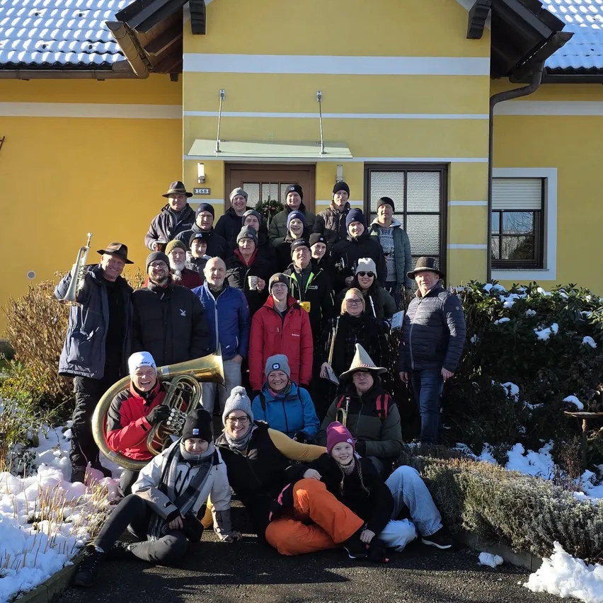 Eine Gruppe von Menschen in Winterkleidung, einige mit Mützen und Brillen, stehen und sitzen vor einem gelben Haus, das von Schnee bedeckt ist. Musikinstrumente sind vorhanden, und das Wetter ist kalt.