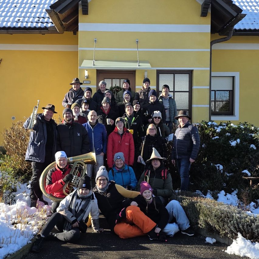 Eine Gruppe von Menschen in Winterkleidung, einige mit Mützen und Brillen, stehen und sitzen vor einem gelben Haus, das von Schnee bedeckt ist. Musikinstrumente sind vorhanden, und das Wetter ist kalt.