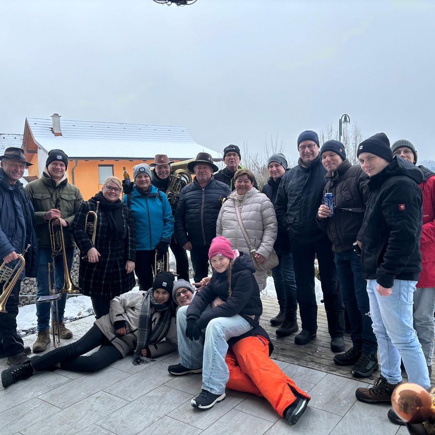 Eine Gruppe von Menschen in Winterkleidung, einige mit Musikinstrumenten, posiert für ein Foto auf einem gefliesten Boden vor einem Gebäude mit Schnee auf dem Dach.