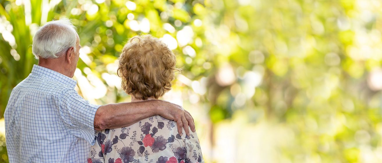 Eine ältere Frau mit kurzen, lockigen Haaren steht in einem Garten, wobei die Hand eines Mannes auf ihrer Schulter ruht. Der Hintergrund ist verschwommen mit grünen Blättern.