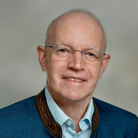Ein Mann mit Brille und einem blauen Pullover mit brauner Einfassung lächelt und schaut direkt in die Kamera.